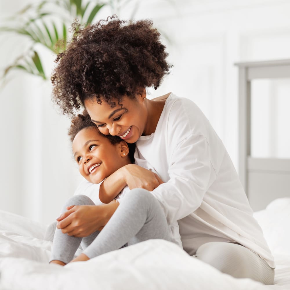 A mother is smiling while hugging her young child. They are both sitting on a bed. 